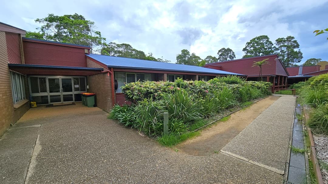 A shaded area with tiered seating extending to the right. Blue plastic chairs are positioned alongside with signage. To the left, another shade structure adjoins a low-rise building with a brown brick facade bearing the name "Merrin Lodge". The roofing structure features corrugated sheets and metal beams. Landscape ornamental plants occupy the front and sides of the building, with trees surrounding the area.