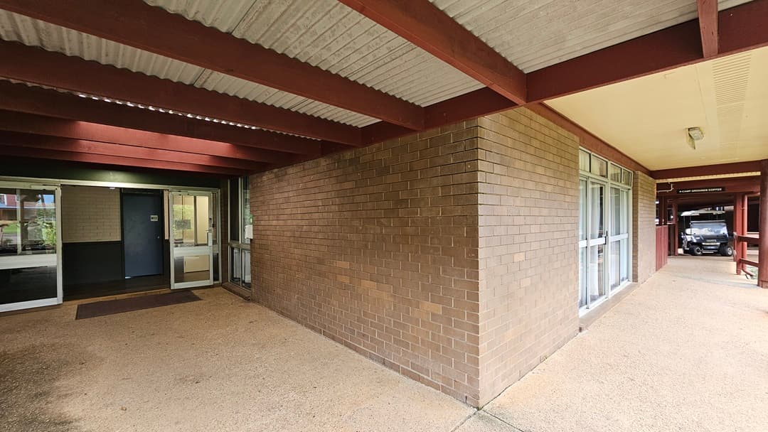 To the left, an outward-opening glass door equipped with matting. The walls have brown brick on the outside and white on the inside. The ceiling has exposed metal beams with a corrugated roof. On the right, a small vehicle is parked.