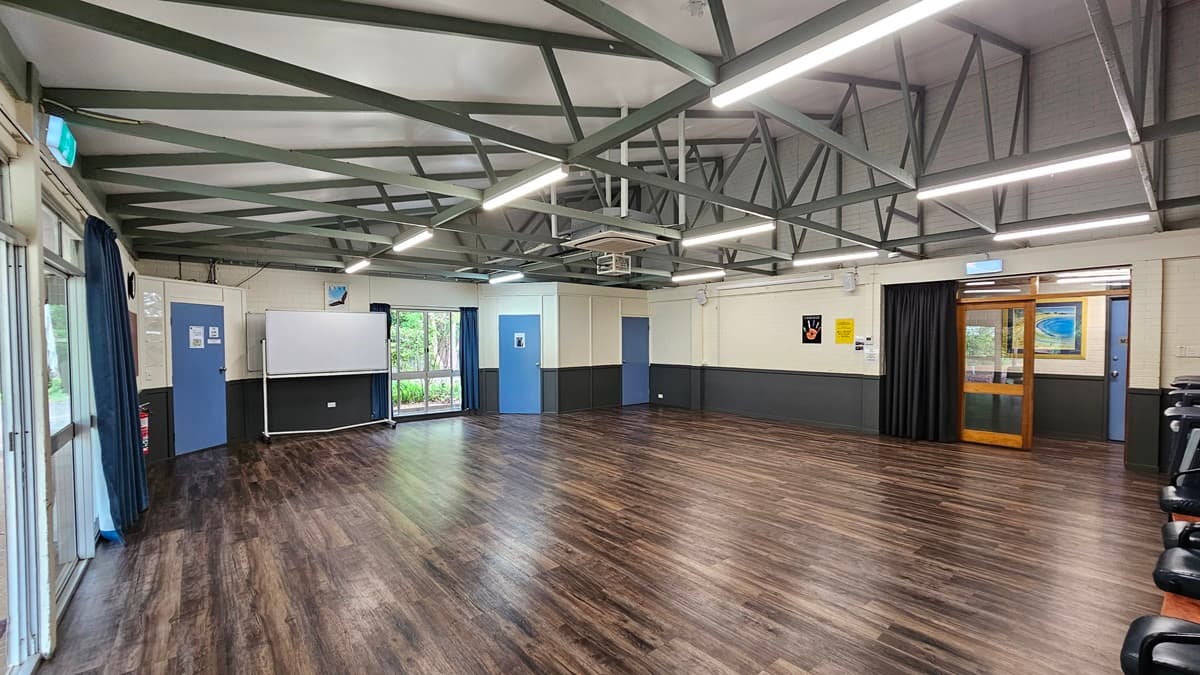 A spacious open area with a brown timber floor. The ceiling features white with exposed metal beams, fluorescent lights, air conditioning, and a projector machine. Glass windows have blue curtains rolled to the sides, next to a movable whiteboard. Blue doors and walls are equipped with posters. To the right is a glass door leading to corridors. Black chairs are arranged against the right wall.