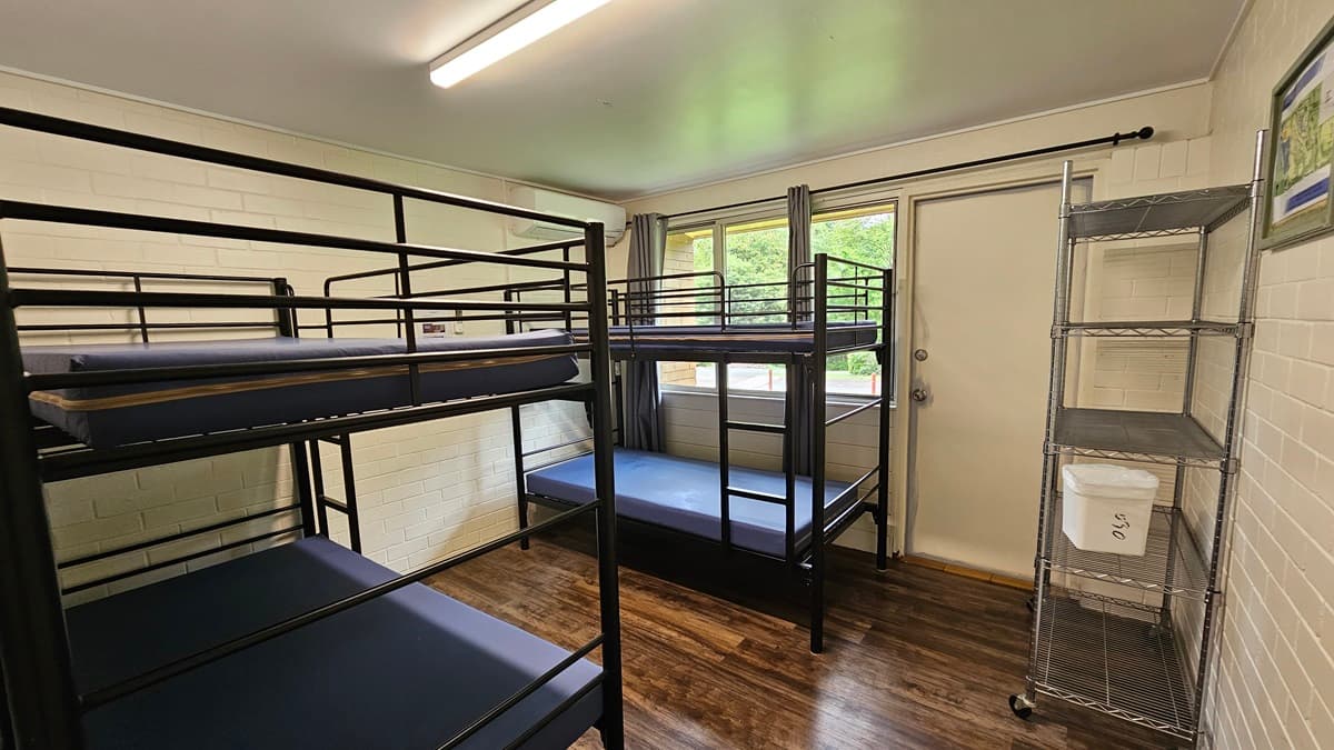A room equipped with metal bunk beds, each with mattresses, positioned against the wall. On the right is a door beside a glass window with grey curtains rolled to the sides. Adjacent to it is a 5-tier movable stainless display rack against the wall. The walls are white with a picture frame decoration. The ceiling is white with fluorescent lights, and the floor is brown timber.