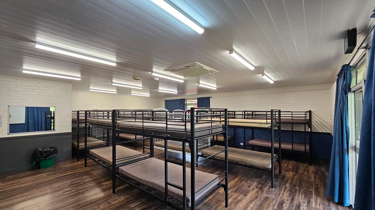 A room equipped with metal bunk beds, each with mattresses. On the right is a door beside a glass window with blue curtains rolled to the sides. The walls are white bricks with a mirror on the left and a rubbish bin below. The ceiling is white with fluorescent lights and air conditioning, and the floor is brown timber.