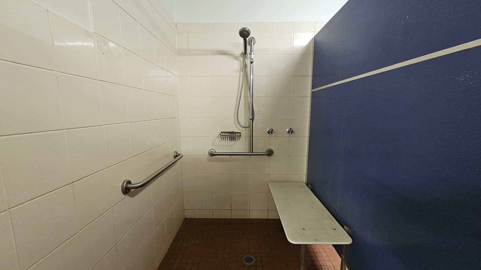 A shower with hand rails and a soap holder mounted on the middle wall. White tiles cover the left and middle walls, with a blue partition wall on the right. Brown tiles adorn the floor. The ceiling is white, and a foldable bench chair is mounted on the right wall.