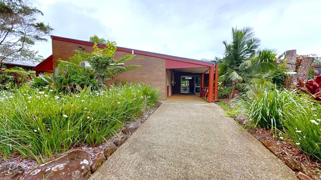 A concrete path leads towards a low-rise building with pitched roofing and a brown brick facade. The path is flanked on both sides by a landscape featuring flowering plants, large rocks, and various trees.