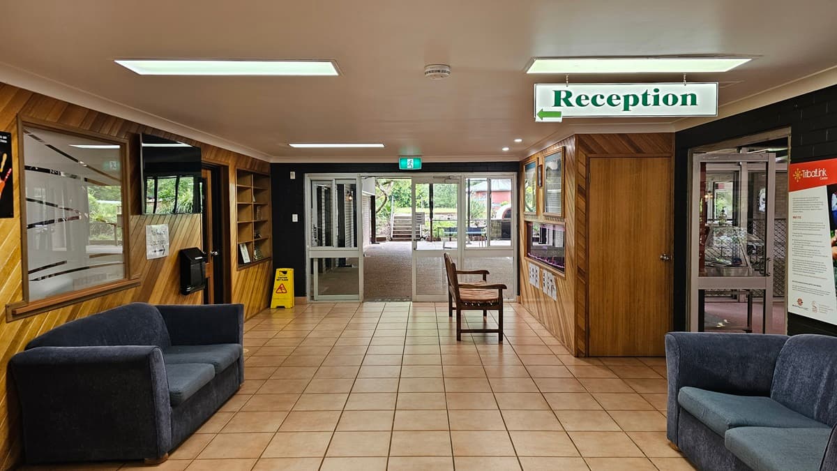 The interior has a low white ceiling with wooden accent walls. Grey sofas are placed on both sides. To the left is a door leading to the reception, where a hand sanitiser and a flatscreen TV are mounted on the wall. On the right, an inward-opening glass door is present. A wooden display cabinet, paintings, and an aquarium is positioned on the right with two chairs in front of it. Various signs are affixed to the wall and suspended from the ceiling.