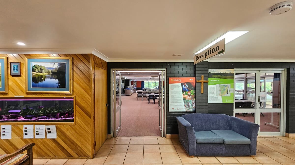 On the left, there's a wooden display cabinet showcasing paintings and an aquarium, with two chairs positioned in front. To its right, an inward-opening glass door leads to the dining area, furnished with plastic chairs and tables. The flooring changes from cream outside to printed brown inside. Various signs are displayed on the walls and suspended from the ceiling.