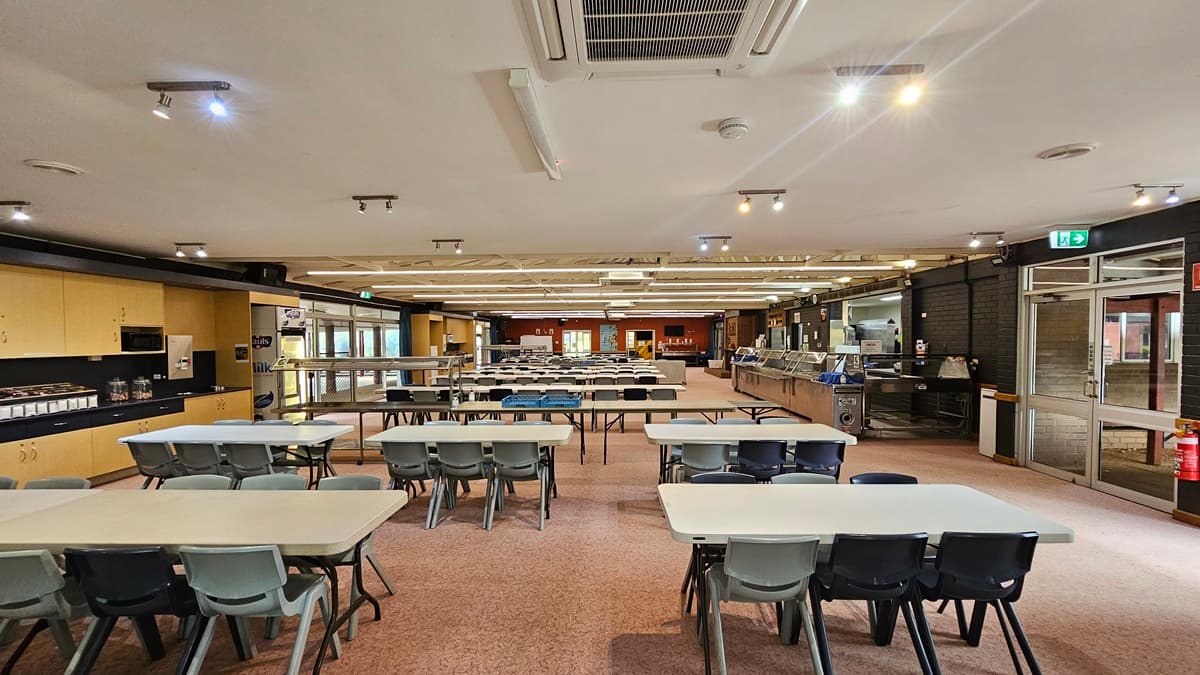 In the middle of the space, rows of tables and chairs are arranged. To the left, there is a cupboard filled with utensils. To the right is a section designated for food display. The low ceiling is white, equipped with LED and fluorescent lights, as well as air conditioning. The floor is adorned with printed brown tiles. On the right, a manual glass door serves as the exit, with a fire extinguisher and an exit sign mounted on the nearby wall.