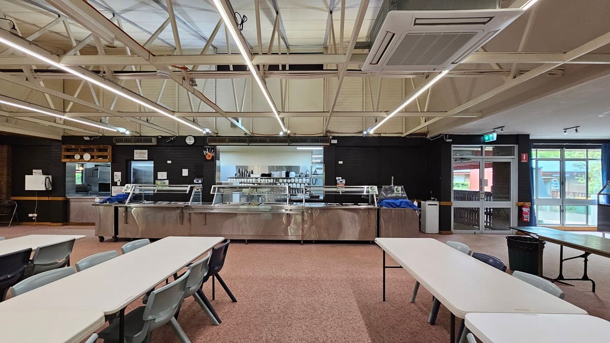 White plastic tables and chairs are neatly arranged in rows. A section for displaying food and utensils is visible in the background. The ceiling has exposed metal beams with fluorescent lights and air conditioning vents. The floor is covered with printed brown tiles. On the right, there's a manual glass door serving as an exit, with a fire extinguisher and an exit sign mounted on the nearby wall.