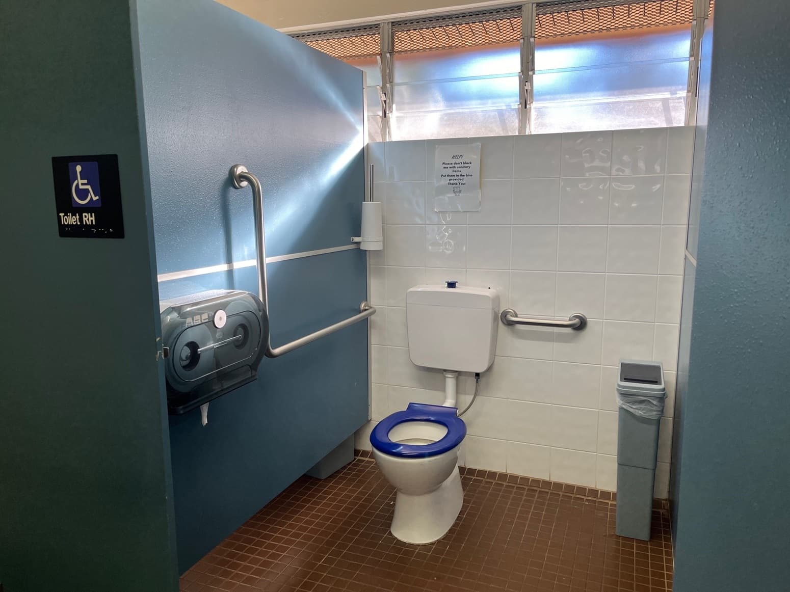 Inside the accessible toilet. A toilet bowl with handrails is in the left corner. A tissue roll dispenser is fixed to the blue wall. A toilet brush is mounted to the upper right blue wall. The back wall features white tiles and a glass window, while the floor is tiled brown. A reminder is affixed to the wall.