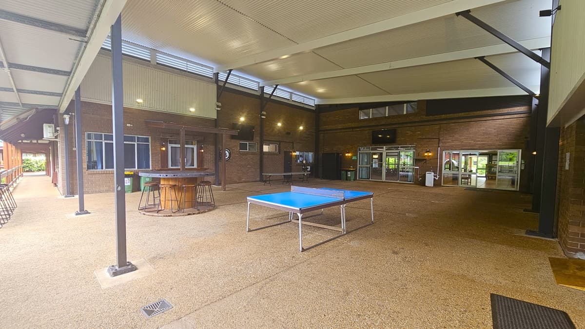 A spacious area with a round table and stools on the left, and a ping pong table in the middle-right. Brown brick walls and a cement floor with a sandy texture. White ceiling with exposed metal beams and corrugated roofing. A black mat is on the right. Flat-screen TVs are mounted on the upper middle and left walls, while white rubbish bins are below a window on the left.