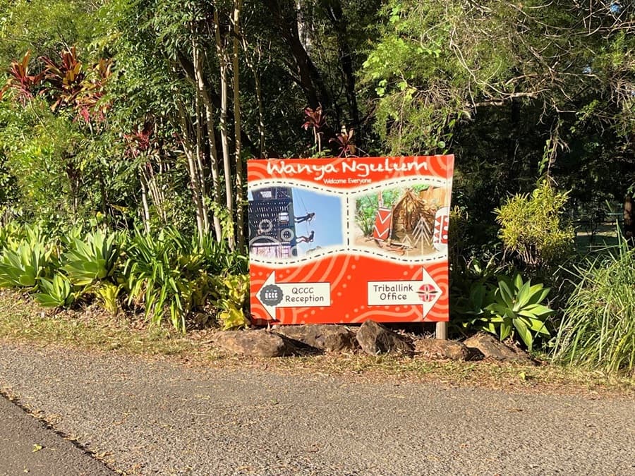 A concrete pathway slopes downward. On the right, a sign reading "TribalLink" is affixed to a wooden post alongside another signage. The path is bordered by plants, native grass, and trees. In the background, the roof of a facility is visible.