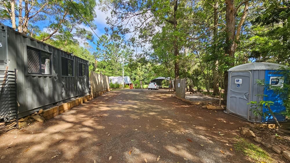 On the left stands a portable building with corrugated metal walls painted grey. Adjacent is a portable toilet, shaded by a tall tree. Further ahead is a dome-shaped structure and a white car. The area is surrounded by trees.