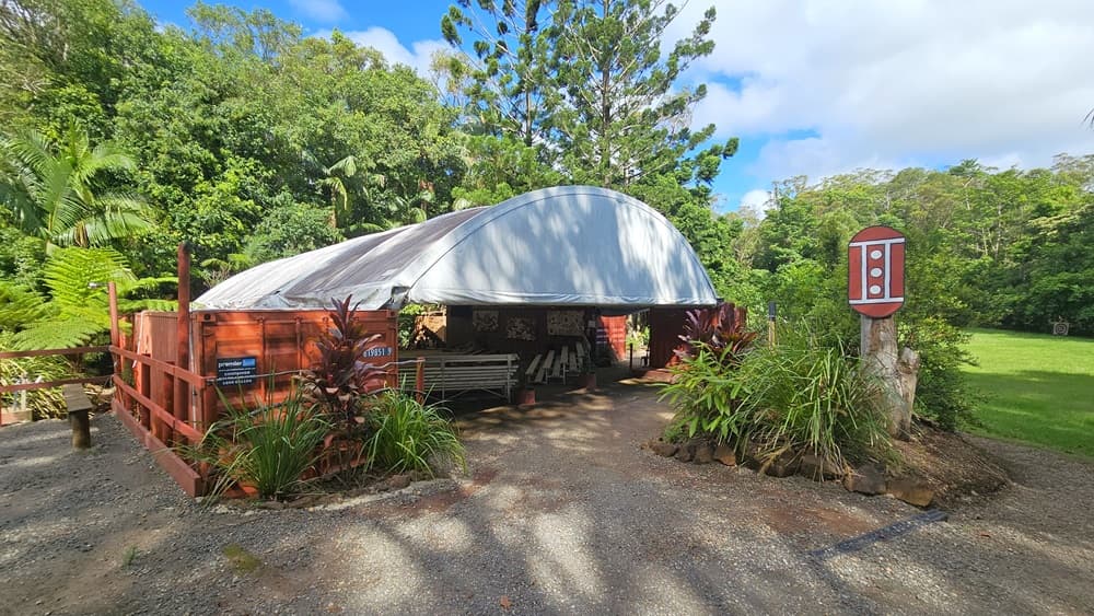 In the centre of a slightly elevated unpaved area stands a dome-shaped tent surrounded by a metal fence. Plants border the front. A signage resembling a traffic light is mounted on a tree trunk. Inside the tent are bench chairs and paintings on the wall. Trees and grass border the right side.