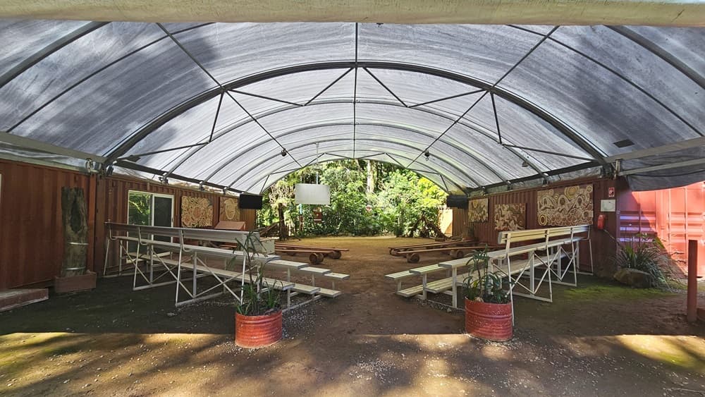 A dome-shaped tent with shade cloth roofing and unpaved ground. Inside, metal beams follow the contour. Tiered seating and improvised bench chairs are arranged on both sides with space in between. The red corrugated wall displays tribal paintings on the right. Indoor plants are placed in front and on the sides, with more visible behind. Flat-screen TVs and projection boards are also in place.