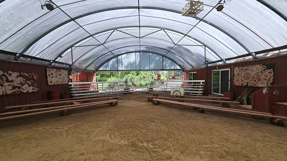 A dome-shaped tent with shade cloth roofing and unpaved ground. Inside, metal beams follow the contour. Tiered seating and improvised bench chairs are arranged on both sides with space in between. The red corrugated walls display tribal paintings. Indoor plants are placed in front and on the sides, with more visible behind. Flat-screen TVs and projection boards are also in place.