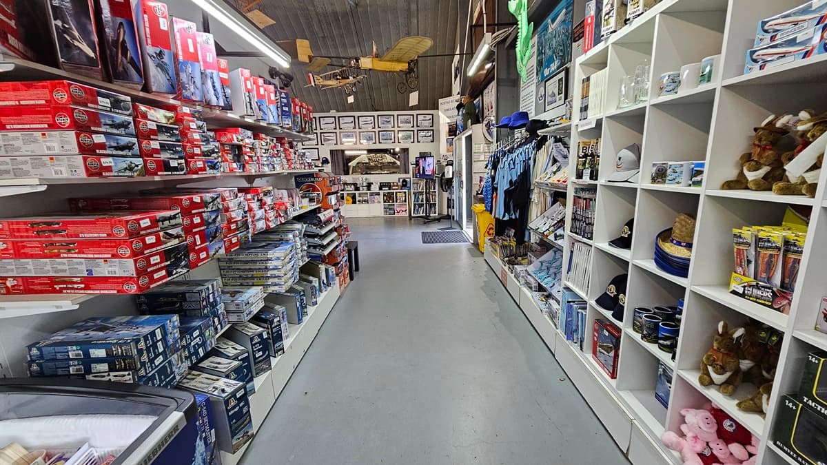 On the left, a reception counter features a welcome sign, informational brochures, and aviation merchandise. A person stands behind the counter in front of a wall decorated with posters and a large circular sign. On the right, various large aircraft models and exhibits are displayed, with a black aircraft in a prominent position. The floor is grey concrete, and the ceiling is metallic with geometric structures, along with several aircraft models suspended above.