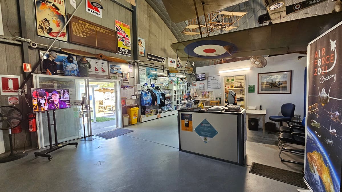 On the left, posters and information boards adorn the wall, beside an entrance with glass doors and a flatscreen showing images. To the right, a customer service desk features a computer terminal and informational signage. Display shelves and merchandise are visible inside. The floor is a polished grey surface, and the high ceiling is decorated. Additionally, chairs and aviation-themed posters are on the right, with a model aeroplane suspended overhead.