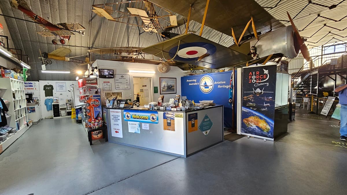 On the left, there is a retail area with shelves displaying various items, clothing racks, and a counter with brochures and merchandise. On the right, there is a large aviation-themed display with a prominent biplane suspended from the ceiling. In the background, walls decorated with aviation posters, memorabilia, and information panels. The foreground features a grey concrete floor and a high, corrugated metal ceiling with additional model planes hanging from it.