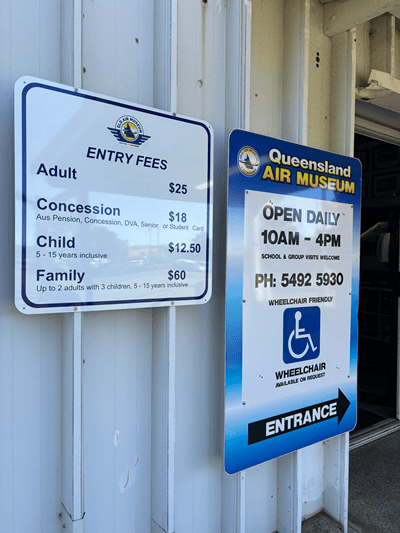 Signage at the entrance of the Queensland Air Museum. On the left side, there is a sign detailing the entry fees. Both signs are mounted on the exterior wall of a building, likely near the entrance door to the museum.
