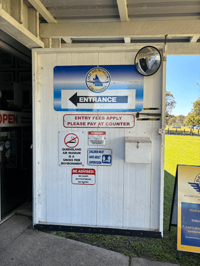Entrance to the facility. Several signs providing information and instructions are affixed to a white wooden door left open. The overall setup suggests the entrance to a regulated public facility, likely prioritizing safety and compliance with specific rules. Green grass fills the backdrop.