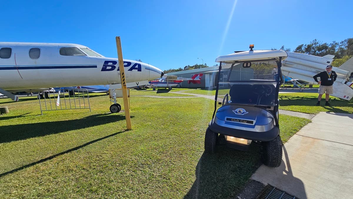 A grey golf cart with a white roof is situated on the left side. The golf cart has black seats and is parked on a concrete surface adjacent to a grassy area. Towards the right, a white aircraft with the registration number BP stands in front of a hangar. In the background, several more aircrafts are visible on display on the grass. The foreground includes a concrete path and the grass appears well-maintained.