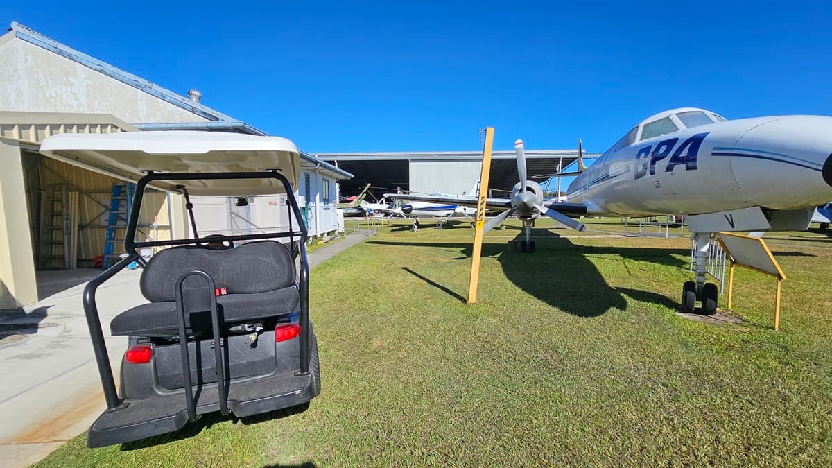 On the left, a golf cart is parked on a concrete path beside a beige, metallic building. In the centre and towards the background, multiple light aircraft are visible, some partially within a large open hangar. On the right, a white jet aeroplane marked "CPA" on its fuselage is positioned on green grass, supported by a metal railing. The foreground includes well-maintained grass and signage.
