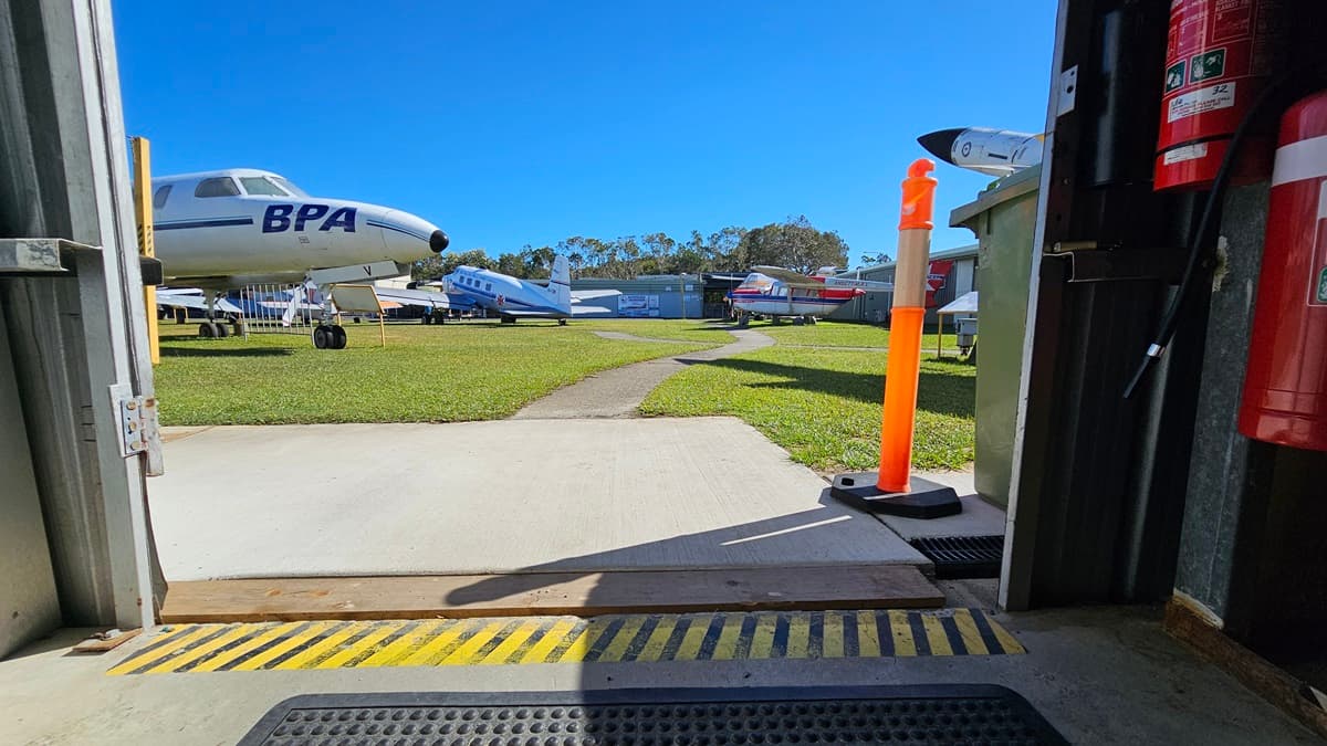 A view from inside a hangar with the doors open. On the left, there are several parked aircraft, including one with 'BPA' written on its fuselage. In the middle, there is a concrete path leading to a grassy field with more aircraft in the background. On the right, an orange traffic cone stands near the doorway. The hangar floor is grey concrete with a ribbed mat and a yellow-black hazard strip at the entrance. The ceiling and rest of the hangar interior are not visible.