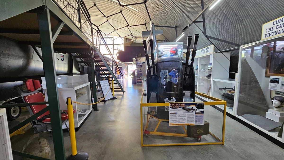 On the left, there is a display case filled with various memorabilia and a small information kiosk. Above it hangs the front section of an aircraft. In the centre, there is a black and silver aircraft surrounded by safety barriers, with a metal staircase with handrails providing access to its cockpit. In the background, on the right, additional aircraft parts and displays are visible. The floor is concrete, and the ceiling is an arched, ribbed structure made of metal.