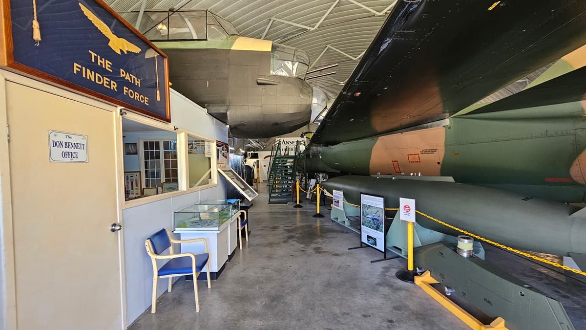 On the left, a dark blue sign stating "The Pathfinder Force" is next to a white door with a small window and another sign. A blue chair and a small desk rest against the wall. On the right, a green and brown military aircraft is encircled by display boards and rope bollards. In the background, various exhibits and information panels are visible. The floor is grey concrete, and the ceiling features a corrugated metal design with overhead lighting.