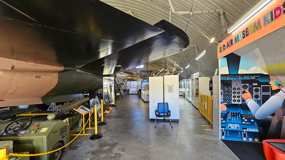 On the left, there is a large aircraft with its wing extending overhead, and yellow barriers along the walk path. Beneath the wing, there are various machines and equipment. On the right, there is an interactive exhibit for kids with colourful graphics and control panels. In the background, the space opens to more displays and information boards. The floor is concrete, and the ceiling is curved with visible ribbed metal panels and lighting fixtures.