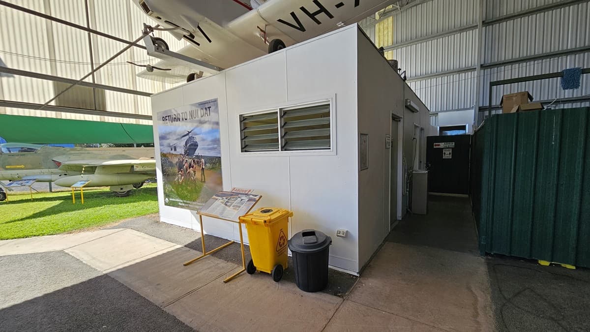 On the left, a light green building features a "Toilet" sign with an arrow pointing downwards. In the centre, a passage with a corrugated overhead sheet leads to an aircraft exhibit, marked by an "Entry" sign above. On the right, a military helicopter is displayed, its rotor blade visible, on a gravel surface. The foreground consists of gravel, grass, and a concrete pathway, while the ceiling is made of corrugated metal with exposed beams.