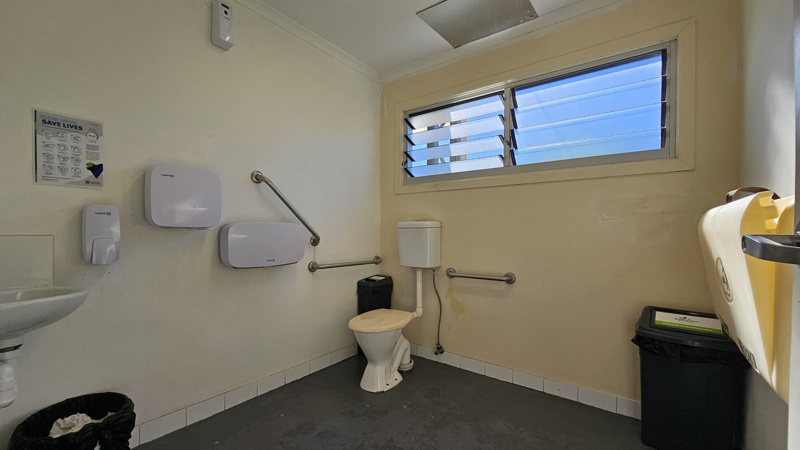 An accessible bathroom includes a toilet with hand grabs in the corner. To the left wall, a tissue dispenser, hand dryer, sink, and liquid soap dispenser are mounted. A louvre window is central. The floor is tiled in grey, while the walls and ceiling are painted cream. A black rubbish bin and a reminder poster are also visible.