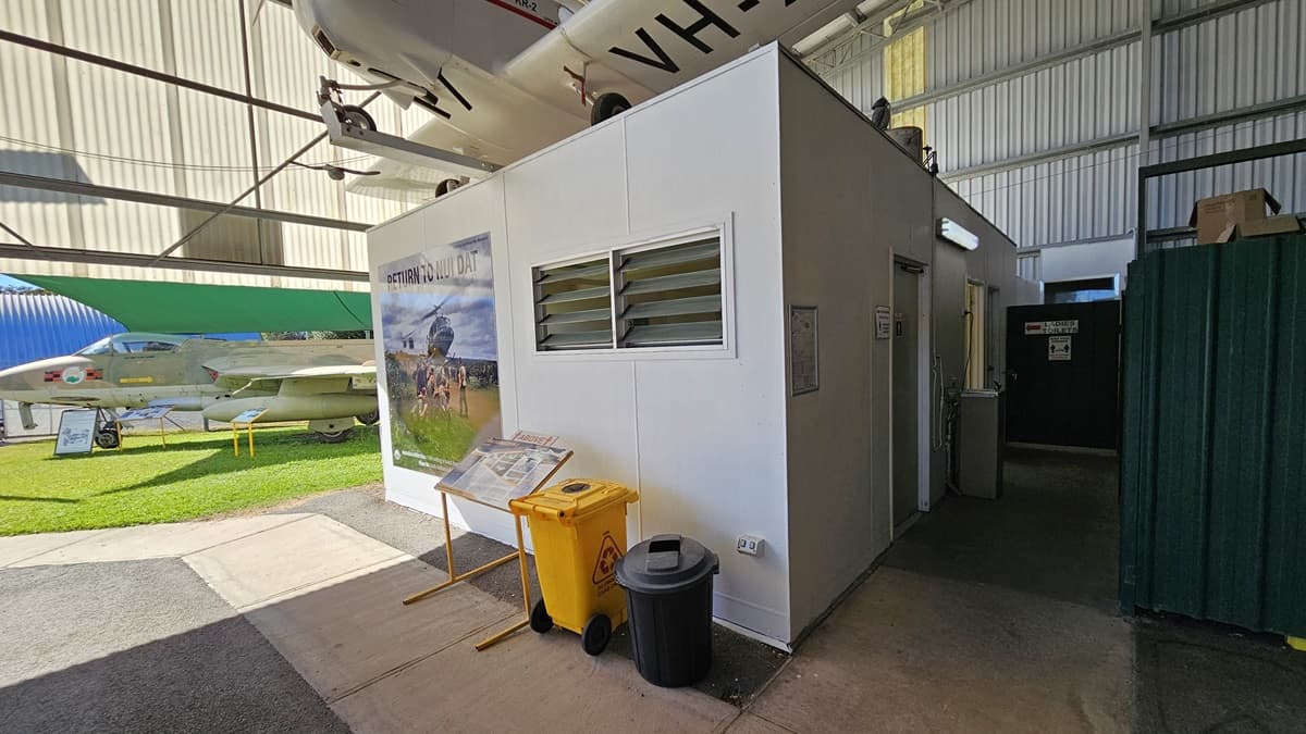 On the left, an area of grass hosts two parked aircraft. An information board and two bins, one yellow and one black, are positioned on concrete against a white wall with an illustrated poster. The right features a white cubicle-like structure with a louvred window and door. The foreground is a mix of concrete and grass; the interior floor is light grey. The background shows a large green wall and a building with metallic cladding, with structural beams visible.