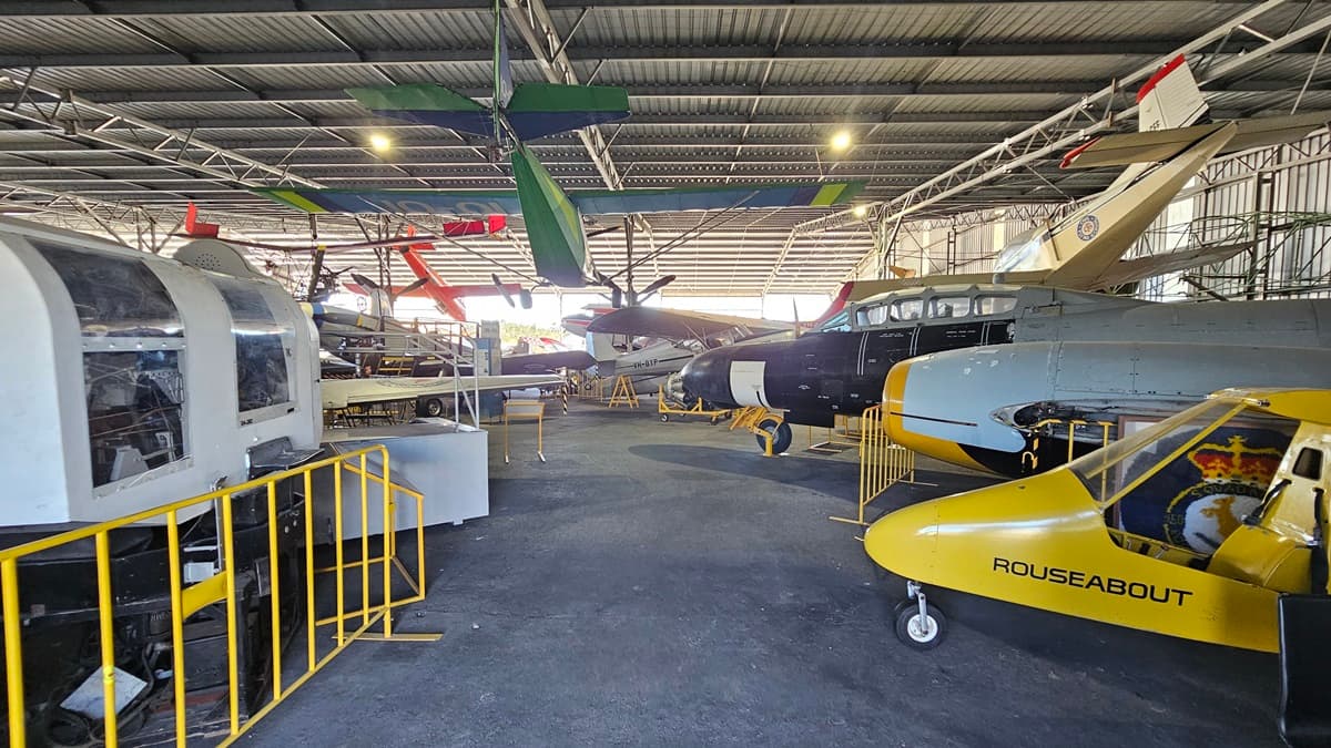 Aircraft hangar with several planes. On the left, a light grey aircraft cockpit with yellow railings around it. In the background, various aircraft with propellers, including green, red, and black ones, are arranged in rows. On the right, a yellow aircraft marked "ROUSEABOUT" is displayed prominently. The floor is concrete, and the ceiling is lined with metal beams and has several light fixtures. There are large airplane models mounted to the ceiling.