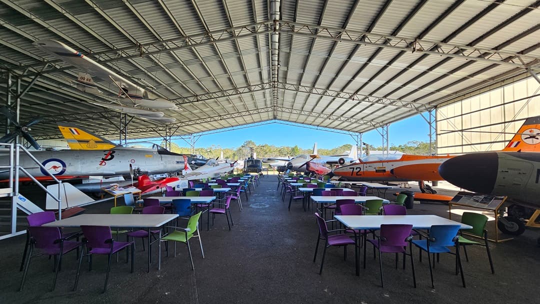 Under a large metal hangar with a corrugated ceiling, several historical aircraft are parked in the background. On the left, there are jet aircraft with distinctive military markings. On the right, more aircraft are visible, one with an orange tail. In the foreground, numerous tables and colourful chairs are arranged on a grey concrete floor, suggesting a dining or event area. The open sides of the hangar reveal trees and greenery outside.