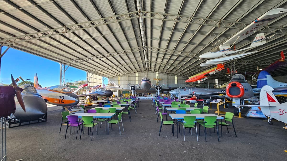 A large open hangar with a corrugated metal ceiling and a concrete floor. In the foreground, there are numerous tables with brightly coloured chairs in purple, green, and white. On the left, vintage aircraft are displayed, including a plane with the number 12. On the right, more vintage aircraft are exhibited. In the background, more aircraft are suspended from the ceiling. The far end of the hangar reveals additional planes and vehicles.