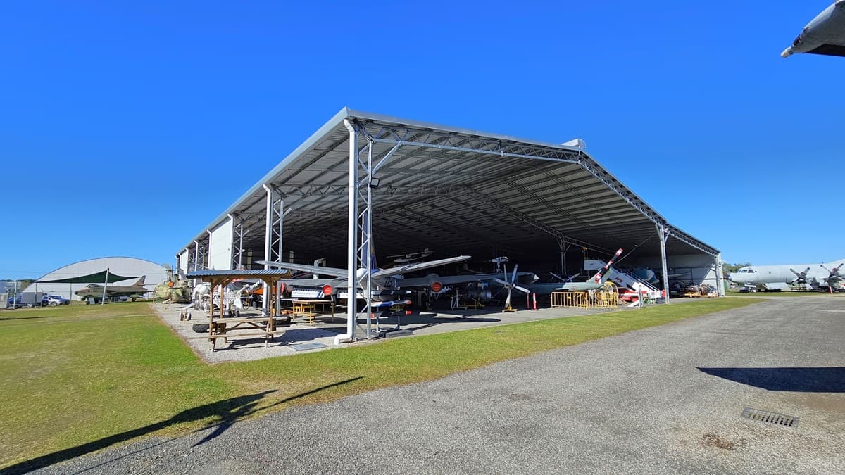 Several aircraft are parked under a large, open-sided hangar with a corrugated metal roof. Wooden picnic tables and benches are placed on a nearby grassy area. To the right, additional aircraft are under the hangar, with an open, paved area extending to the foreground. The hangar has a metal roof supported by metal beams and a concrete floor. In the background, more hangars and aircraft are visible. The ground comprises grass and gravel surfaces transitioning to a paved path.
