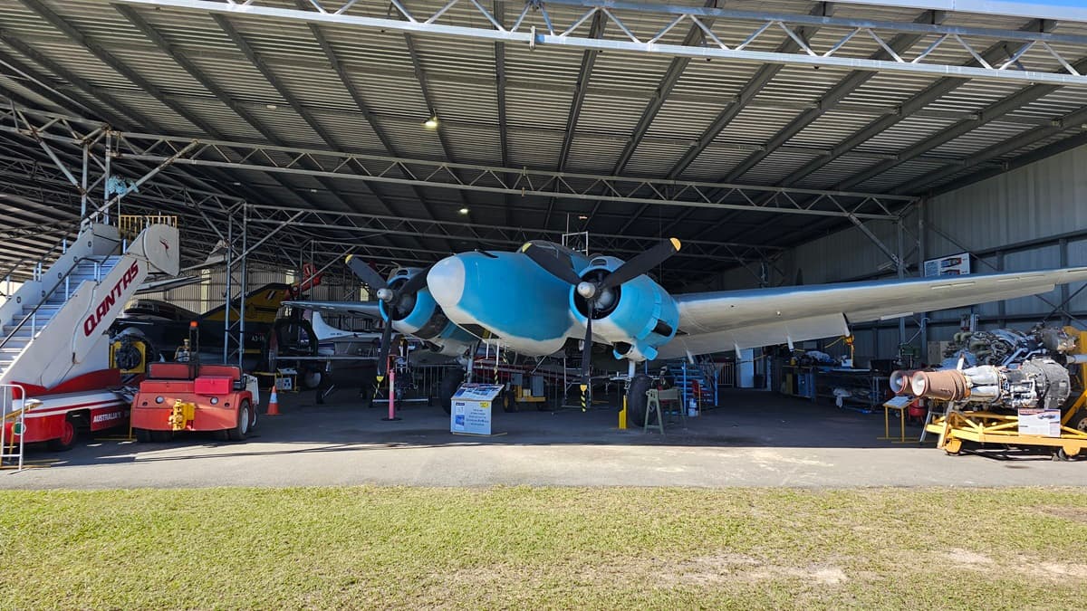 A blue and white aircraft is centred beneath a large metal hangar. To the left, a large red and white aircraft stairway with "QANTAS" railings is surrounded by industrial tools and equipment. To the right, additional machinery and various aeroplane components are visible, along with part of another aircraft. The floor is concrete, edged with grass in the immediate foreground. The ceiling is made of corrugated metal panels with overhead lighting. Grass covers the foreground.