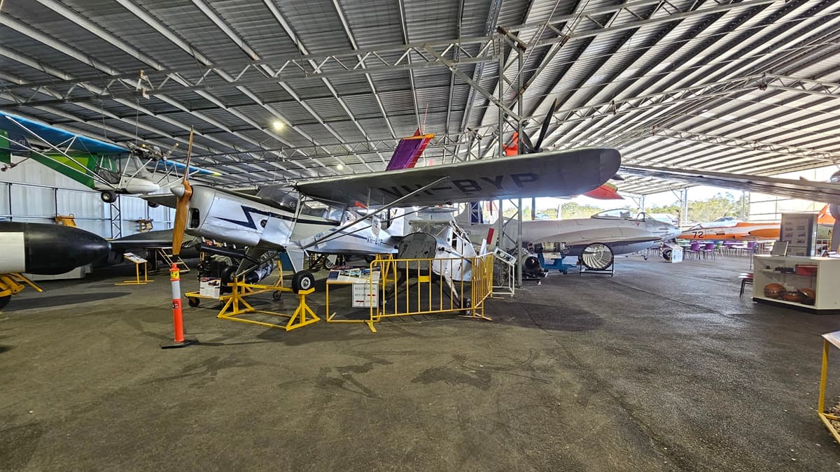 On the left, a vintage aircraft with a distinctive propeller is secured by yellow railings and an orange bollard, with a colourful biplane suspended overhead. In the centre, another classic aeroplane is parked behind yellow barriers, its identification code "VH-BVP" visible. The foreground shows a smooth, dark concrete floor. The ceiling features a metallic, corrugated structure with large beams and numerous suspended lights. A white wooden information desk is positioned on the right.