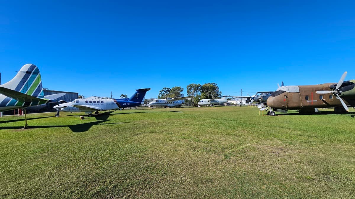 On the left, there are several smaller aeroplanes with white and blue markings. On the right, a large, grey aircraft with four propellers is on display. The foreground consists of a gravel path leading to a grassy area where the planes are positioned. Several other aircraft can be seen scattered in the background, alongside trees and a building. The setting is an open-air plane exhibition area with clear visibility of each aircraft.