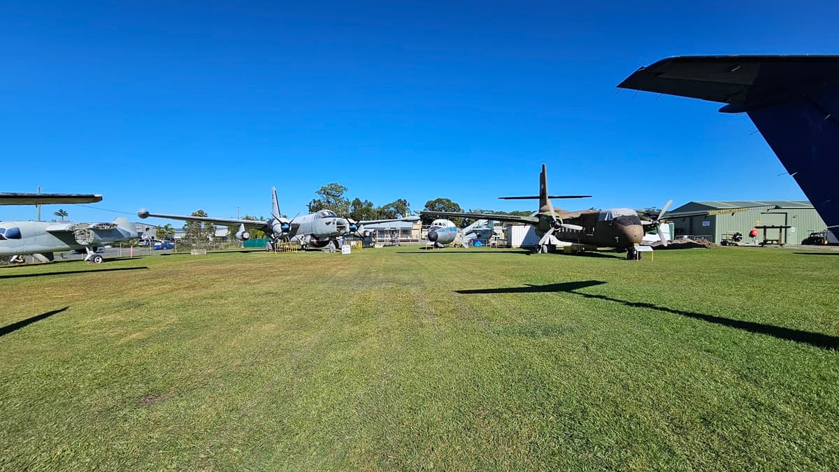 On the left, several aeroplanes with white and blue markings are on display. To the right, a large grey military aircraft with four propellers is featured. The entire area is covered in green grass. In the background, more aircraft, trees, and a building can be seen. A low, green building with a corrugated facade is also on the right.