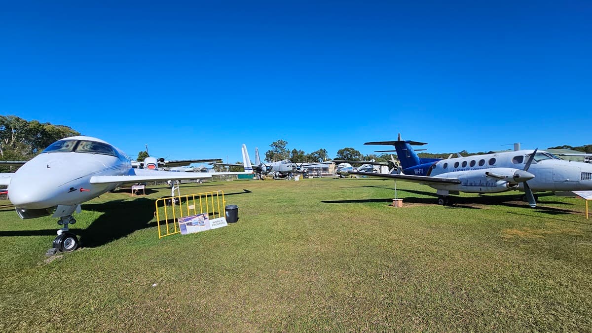 On the left, there is a white aircraft resting on a grassy field. In the middle distance to the left, there is a yellow barrier with a sign attached to it. To the right, another aircraft with a mix of blue and white paint is positioned on the grass. The foreground consists of green grass, and a collection of various planes is visible in the background. Trees and buildings are further back.