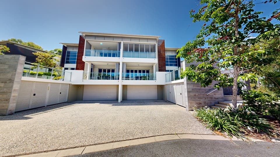 A three-story twin-style building made of concrete and glass, with a spacious front and a garage. Steps with handrails on the right lead to the upper deck. Tall trees and flowering plants adorn the front landscape.