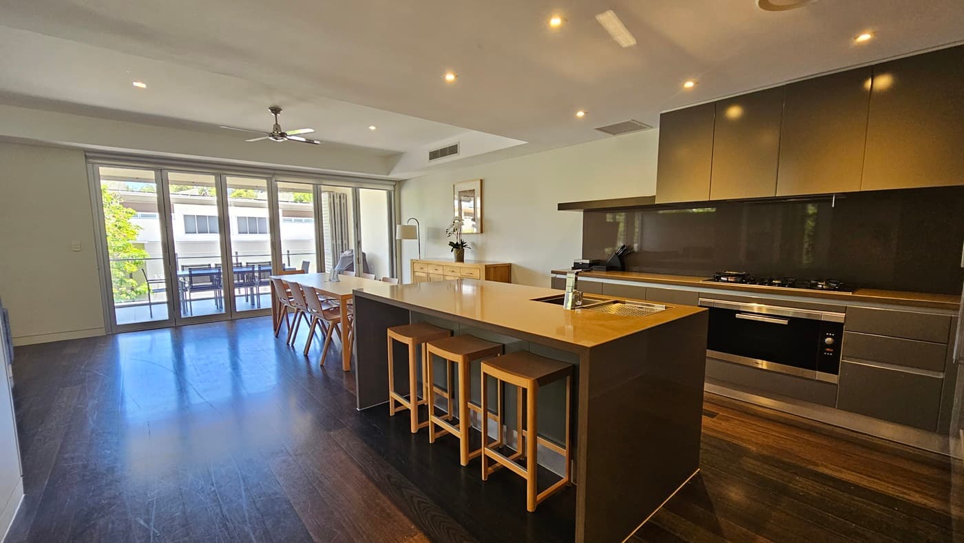 In the kitchen, a central island with a sink, is surrounded by cooking equipment and cabinets. Adjacent is a dining table with a display cabinet nearby. A framed decoration adorns the white wall. Beyond, tables and chairs are visible on the balcony. The floor is polished timber, and the wall and ceiling are white.