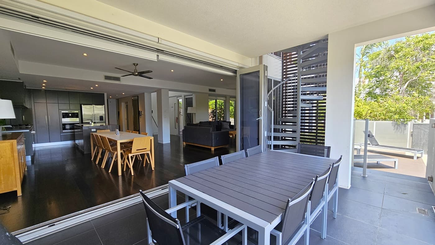 To the right, movable table and chairs are arranged. A spiral staircase leads to the upper deck, bordered by a glass wall railing. On the left, the kitchen area features timber floors, a wooden dining table and chairs, and a lounge area. A kitchen island, cabinets, equipment, and a large fridge are present. A wooden display cabinet holding a lamp stands on the left. The white ceiling is adorned with a ceiling fan and LED lights.