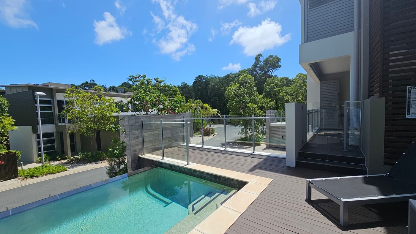 A plunge pool with steps and black sun loungers on a timber deck. Glass railing secures the area. The building facade, a blend of concrete, glass, and timber, is on the right. Trees, plants, and other villas are visible below.
