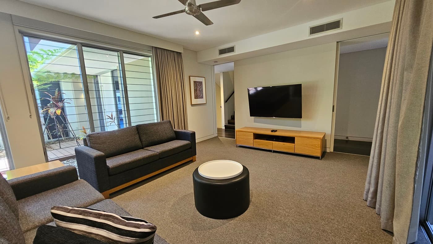 The lounge area consists of sofas arranged around a round center table facing a large flatscreen TV on a TV stand. Floor-to-ceiling grey curtains hang on both sides. A glass wall on the left provides a view of plants and trees outside. A framed decoration hangs on the left wall. Stairs with handrails lead upwards on the left. The floor has grey carpet, and the ceiling is white with a mounted ceiling fan.