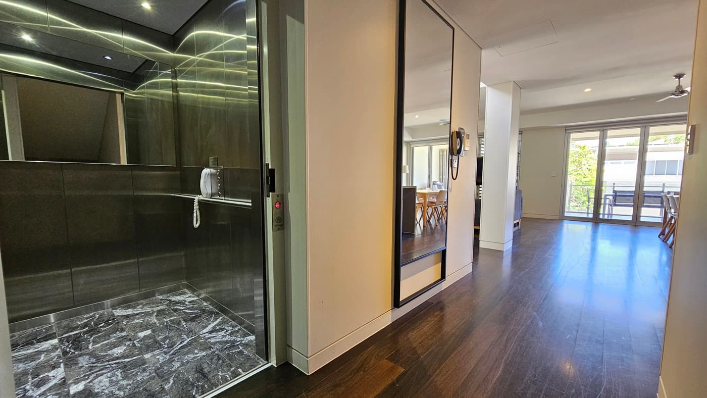 To the left is a lift with a mirror and phone. Adjacent is the entrance to the suite. A large mirror hangs on the left wall, next to a landline phone. To the right, a table and chairs are visible. The floor has polished timber, while the wall and ceiling are white, with a ceiling fan.