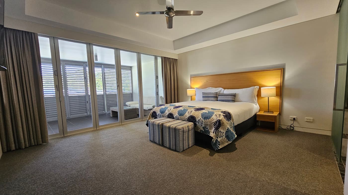 On the right is a king-size bed with printed linens and a wooden headboard, flanked by matching bedside tables and lampshades. To the left is a glass partition that, when open, grants access to the secured balcony. Floor-to-ceiling grey curtains are drawn to the left. The floor is carpeted in grey, while the ceiling features a cove design with pin lights and a ceiling fan, and the walls are white.