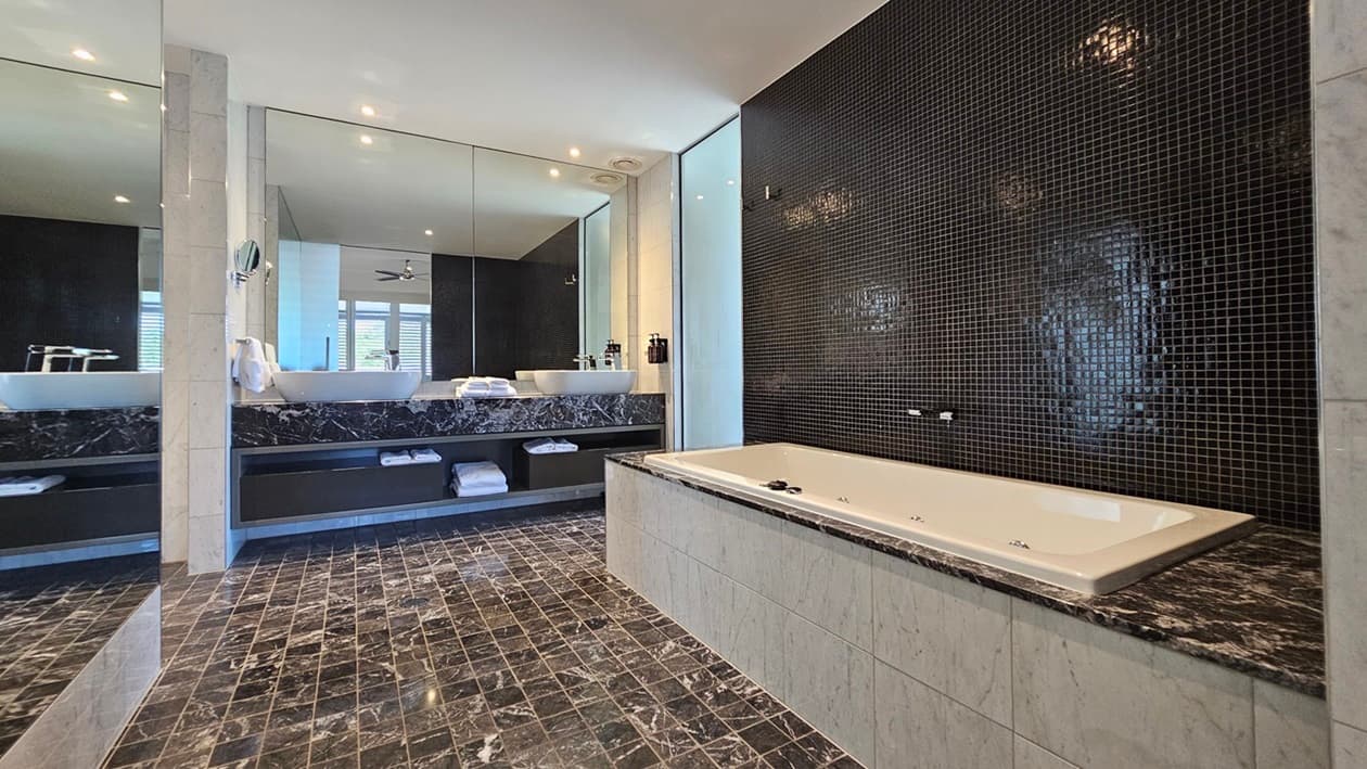 A spacious bathroom with a tub on the right. The right wall features small black tiles. The floor has printed black tiles matching the lavatory. In the middle, a large mirror sits above two hand basins with a towel organizer underneath. Liquid soap dispensers are mounted to the wall. Another large mirror is placed on the left, facing the tub. The ceiling is all white with LED lights and a ceiling fan.