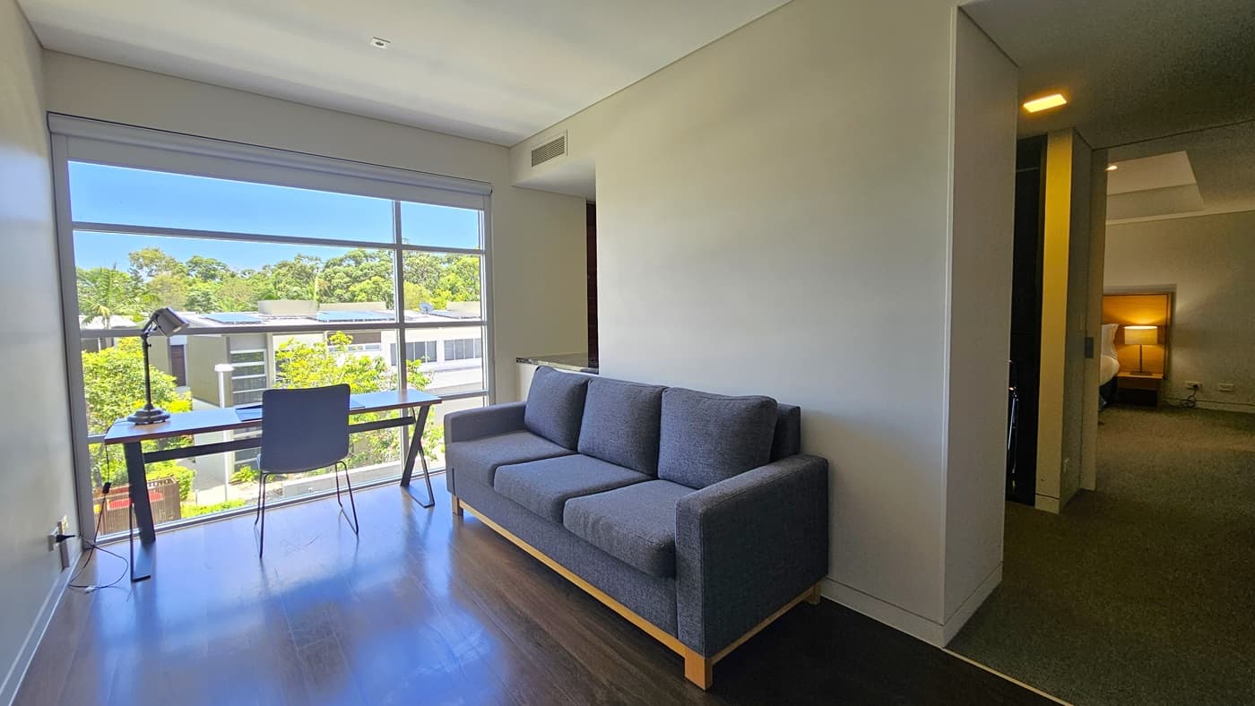 An office table and chair are positioned facing the glass wall, offering a view of the villas and trees. A desk lamp is on top. To the right is a grey sofa. The floor is polished timber. Adjoining the room is the bedroom with grey carpets, white walls, and a white ceiling with LED lights.
